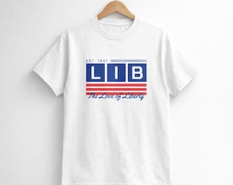 Op de afbeelding: Wit T-shirt met een patriottisch ontwerp. Het ontwerp toont het woord "LIB" in een blauwe rechthoek, met rode en witte strepen eronder. De tekst "The Love of Liberty" staat onder de strepen en "EST. 1847" erboven.