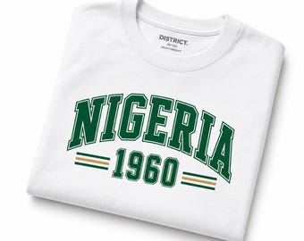 Op de afbeelding: Een wit t-shirt met het woord NIGERIA in een donkergroen collegiaal lettertype, gebogen boven het getal 1960. De tekst wordt geflankeerd door twee horizontale lijnen in groen en beige. Het binnenlabel toont de tekst DISTRICT RE-TEE HEAVYWEIGHT.