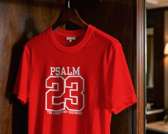 Op de afbeelding: Een rood t-shirt met de tekst "PSALM 23" in witte letters. Onder het nummer 23 staat de tekst "THE LORD IS MY SHEPHERD". Het shirt hangt aan een houten hanger.