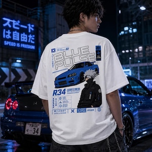 以下が含まれることがあります： 青い車のグラフィック、"BLUE PULSE"の文字、日本語のテキストがプリントされた白いTシャツ。デザインにはキャラクターイラストと"R34"、"SPEED IS FREEDOM"の文字が含まれています。