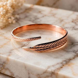 Könnte beinhalten: Ein Armreif aus roségoldfarbenem Metall in Form einer Feder. Das Design zeichnet sich durch einen Wickelstil mit offener Vorderseite aus und zeigt detaillierte, eingravierte Linien, die die Textur einer Feder und ihre feinen Härchen nachahmen.