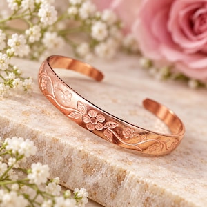 May include: A copper cuff bracelet featuring an embossed floral vine pattern that wraps around the exterior surface. The metal has a warm, reddish-brown metallic finish with a polished texture, showcasing detailed petals and leaves.