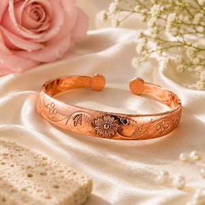Könnte beinhalten: Ein kupferfarbenes Metall-Armband in offener Form mit geprägten Blumen- und Blattmustern auf der Außenseite. Das Armband hat eine polierte Oberfläche und abgerundete Enden, die mit detaillierten botanischen Gravuren verziert sind.