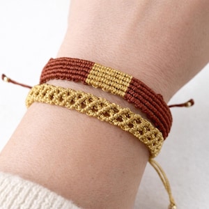 Può includere: Due braccialetti macramè fatti a mano. Uno è una combinazione di marrone e oro, con un motivo rettangolare dorato. L'altro è un braccialetto macramè dorato. Entrambi i braccialetti sono regolabili e annodati.