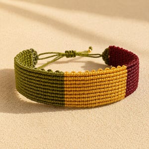 Könnte beinhalten: Ein gewebtes Makramee-Armband mit drei verschiedenen Farbblöcken in Olivgrün, Goldgelb und Dunkelburgunder. Das Armband ist mit einem dichten, strukturierten Knotenmuster gefertigt und verfügt über ein verstellbares Schiebeverschluss-System.