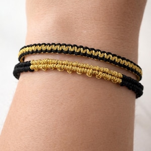 May include: Two macrame bracelets, one above the other, are shown on a wrist. The bracelets are black and gold, with a woven design. The top bracelet has a gold top and black bottom. The bottom bracelet is gold with black on the bottom.