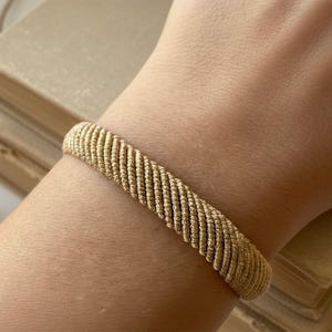 Puede incluir: Una pulsera dorada con un diseño de cuerda trenzada se muestra en un brazo. La pulsera mide aproximadamente 2 cm de ancho. El fondo incluye una pila de libros viejos.