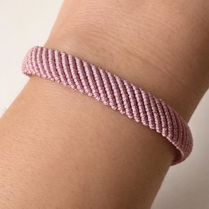 Può includere: Primo piano di un braccialetto rosa realizzato in materiale tessuto. Il braccialetto è avvolto attorno a un polso. Il braccialetto ha un design strutturato e a coste. Il colore è un rosa tenue e smorzato.