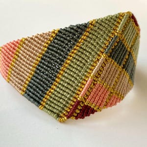 May include: A macrame bracelet with a geometric design. The bracelet combines pink, beige, black, green, and burgundy threads, accented with gold. The bracelet is approximately 5 cm wide.