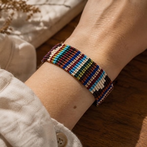 May include: A woven macrame bracelet featuring a repeating pattern of vertical stripes in shades of dark brown, cream, bright blue, and forest green. The bracelet is constructed from thin, tightly knotted threads, creating a flat, wide band.