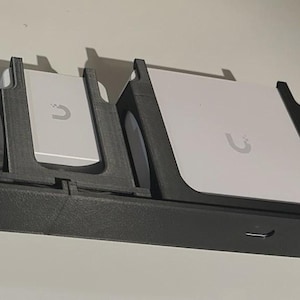 Könnte beinhalten: Ein schwarzer Rack-Mount mit mehreren Schlitzen, die weiße elektronische Geräte halten. Die Geräte haben ein abgerundetes U-förmiges Logo. Das Rack hat mehrere Anschlüsse auf der Vorderseite.