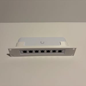 May include: A white network switch with seven Ethernet ports. The device has a rectangular shape with a curved top and a white frame. The brand logo is visible on the top. The device is designed for network connectivity.
