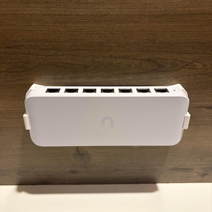 May include: A white network switch with multiple ports, featuring a curved design and a subtle logo. The device is mounted on a wooden surface, showcasing a clean and modern aesthetic. The ports are black and the logo is a white 'C' shape.