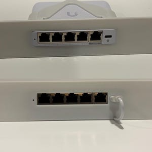 以下が含まれることがあります： 複数のイーサネットポートを備えた2つの白いネットワークスイッチパネル。 上部パネルには5つのポートとUSB-Cポートがあります。 下部パネルにも5つのポートと白いケーブルがあります。 白いデバイスが上部パネルの上にあります。