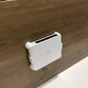 May include: A white network switch with multiple ports is mounted on a wooden surface. The switch has a curved logo and is held in place by white brackets. The wooden surface has a brown wood grain pattern.