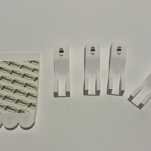 May include: An assortment of white adhesive picture hanging strips and hooks. The strips are rectangular with a repeating logo pattern. The hooks are rectangular with a small, gold-colored knob at the top. The items are arranged on a white surface.