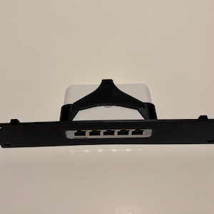 Op de afbeelding: Een zwart netwerkapparaat met vijf poorten is gemonteerd op een zwarte beugel. Het apparaat is wit en heeft een zwarte ondersteuningsstructuur. De beugel heeft montagegaten.