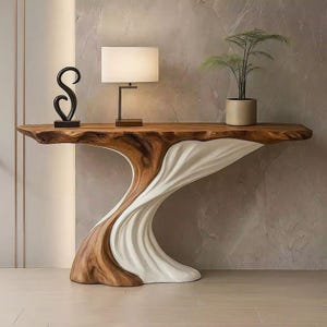 Op de afbeelding: Een moderne console met een houten blad met een natuurlijke rand en een uniek onderstelontwerp. De basis is een combinatie van hout en wit, vloeiend in een golfachtig patroon. Een zwart beeld, een lamp en een plant in een pot staan op de tafel.