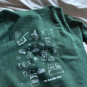 May include: Dark green sweatshirt with white graphic illustrations, including pizza slices, a coffee cup, and the text "THE WALKING DEAD." The sweatshirt features a crew neck and long sleeves.