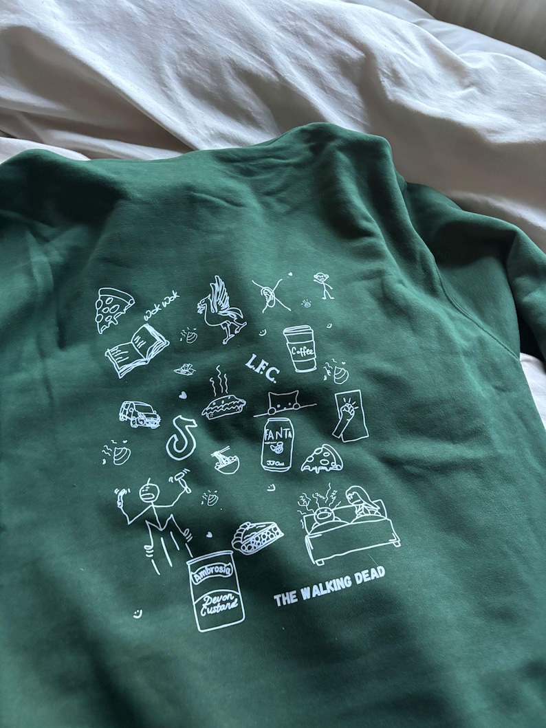 May include: Dark green sweatshirt with white graphic illustrations, including pizza slices, a coffee cup, and the text "THE WALKING DEAD." The sweatshirt features a crew neck and long sleeves.
