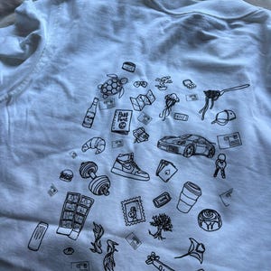 May include: White t-shirt with a black line drawing design. The design includes a turtle, a sports car, a sneaker, a dumbbell, a croissant, a coffee cup, a plane, and other travel and lifestyle-related illustrations.