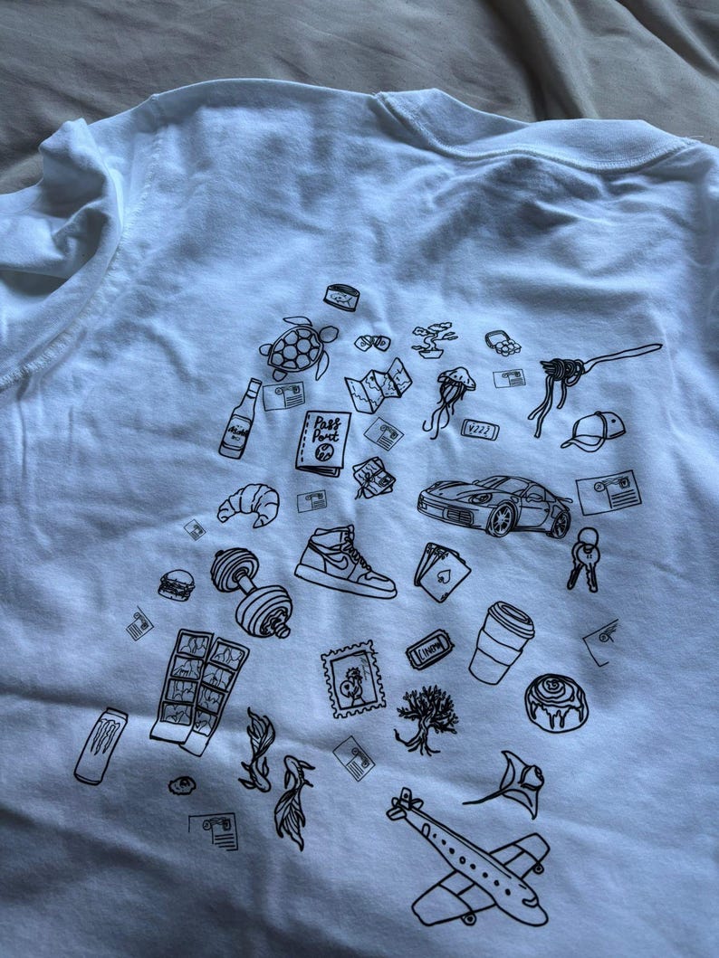 May include: White t-shirt with a black line drawing design. The design includes a turtle, a sports car, a sneaker, a dumbbell, a croissant, a coffee cup, a plane, and other travel and lifestyle-related illustrations.