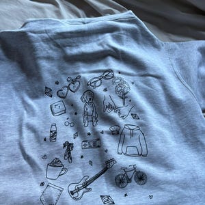 May include: A light gray sweatshirt featuring a collection of black line art illustrations. The drawings include a guitar, bicycle, coffee cup, baby, glasses, flowers, and other small objects. The sweatshirt is made of a soft material.