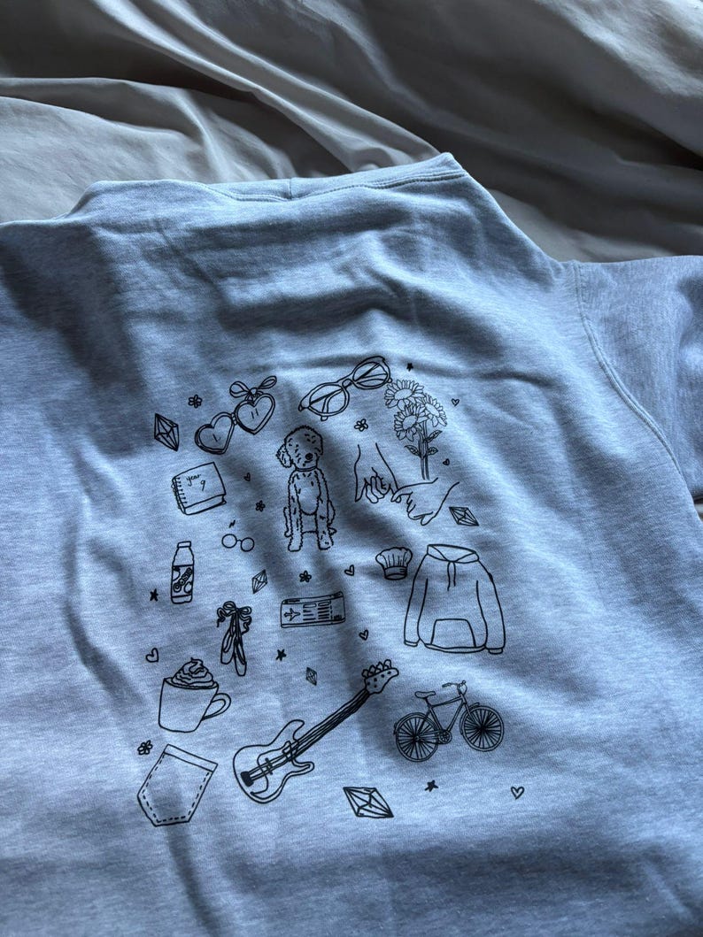 May include: A light gray sweatshirt featuring a collection of black line art illustrations. The drawings include a guitar, bicycle, coffee cup, baby, glasses, flowers, and other small objects. The sweatshirt is made of a soft material.