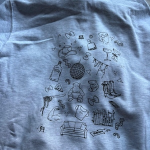 May include: A light gray sweatshirt with a collection of black doodle illustrations. The drawings include a cowboy hat, disco ball, cocktail glasses, a hippo, a couch, and a pair of cowboy boots. The word "KACHOW" is also visible.