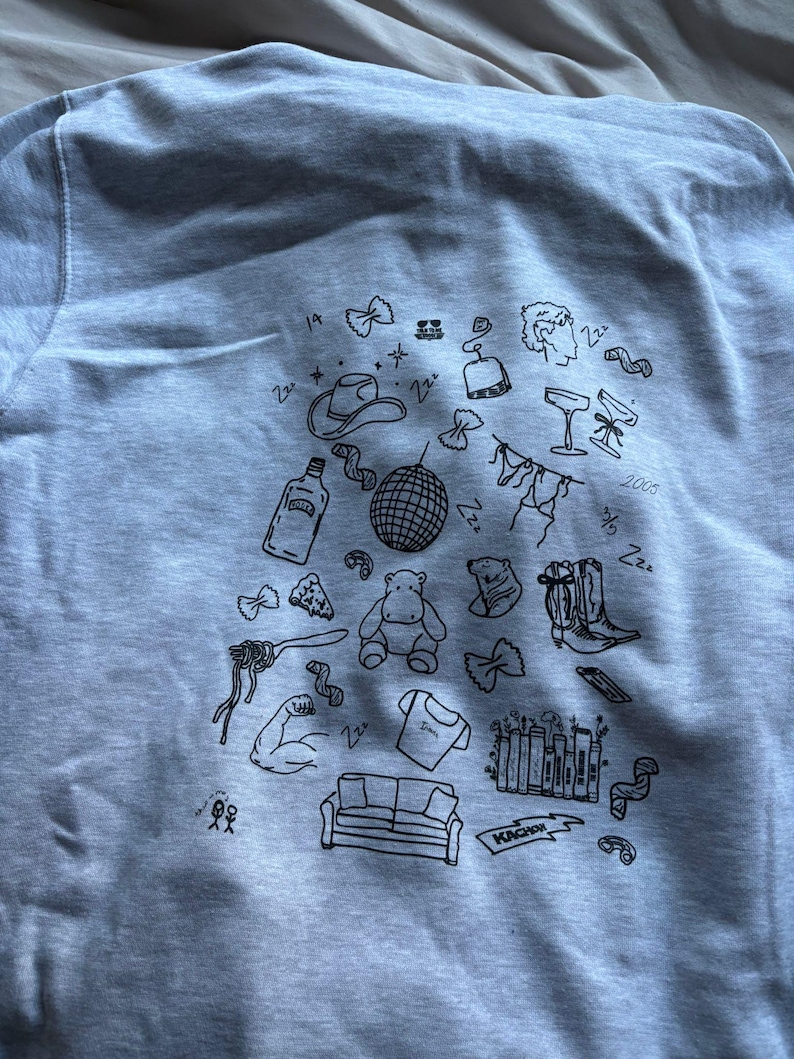 May include: A light gray sweatshirt with a collection of black doodle illustrations. The drawings include a cowboy hat, disco ball, cocktail glasses, a hippo, a couch, and a pair of cowboy boots. The word "KACHOW" is also visible.