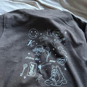 May include: Dark gray sweatshirt with white graphic illustrations. The design includes a smiley face, cat, Fleetwood Mac text, a key, a Coca-Cola can, and other drawings. The sweatshirt is made of a soft material.