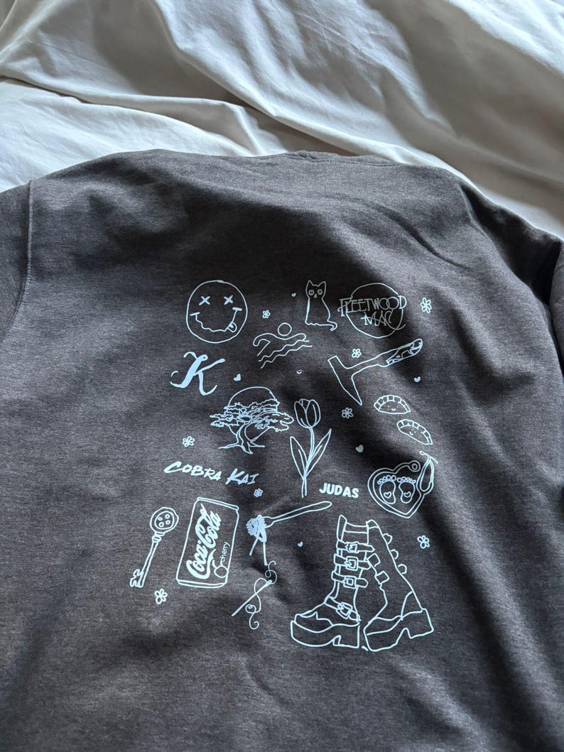 May include: Dark gray sweatshirt with white graphic illustrations. The design includes a smiley face, cat, Fleetwood Mac text, a key, a Coca-Cola can, and other drawings. The sweatshirt is made of a soft material.