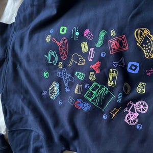 May include: Navy blue hoodie with a colorful graphic design. The design includes various travel-themed illustrations such as a plane, a bicycle, a map, and a shoe. The year "2015" is also printed on the hoodie.