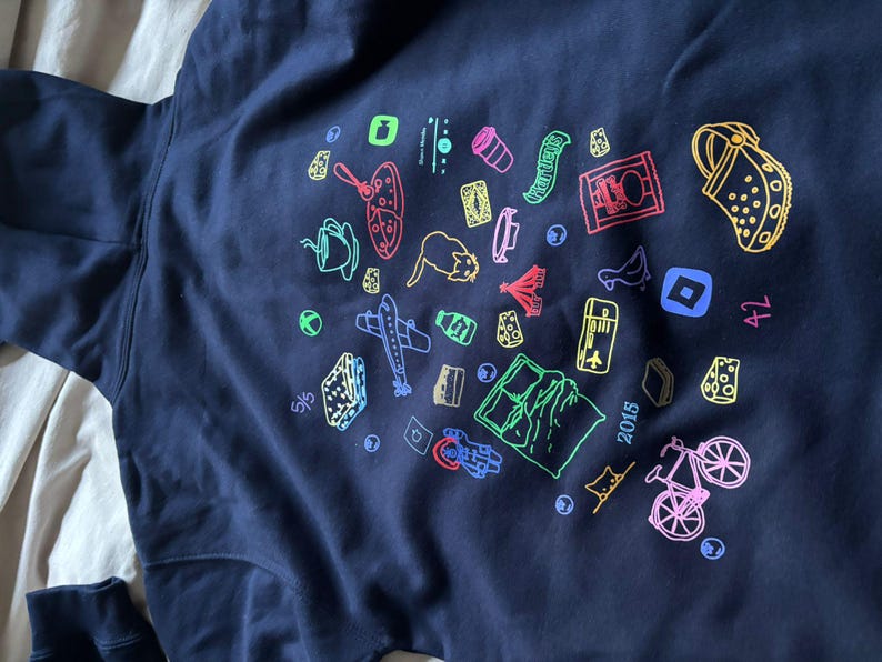 May include: Navy blue hoodie with a colorful graphic design. The design includes various travel-themed illustrations such as a plane, a bicycle, a map, and a shoe. The year "2015" is also printed on the hoodie.