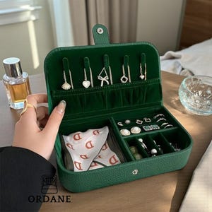 Puede incluir: Una caja de joyería verde abierta, que revela varios compartimentos y espacios para collares y pendientes. La caja contiene anillos, pendientes y una pequeña bolsa. La caja está hecha de un material texturizado.