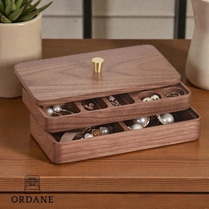 Könnte beinhalten: Eine zweistöckige Schmuckschatulle aus Holz mit einem Klappdeckel und einem goldfarbenen Knopf. Die Schatulle ist mit verschiedenen Schmuckstücken gefüllt, darunter Perlen und Ringe. Die Schatulle ist aus dunkelbraunem Holz gefertigt und steht auf einer Holzoberfläche.