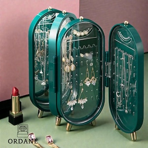 Puede incluir: Un organizador de joyas verde azulado con puertas transparentes, patas doradas y una parte superior dorada. El organizador está abierto, mostrando collares y aretes. Un lápiz labial y joyas están en primer plano. La marca "ORDANE" es visible.