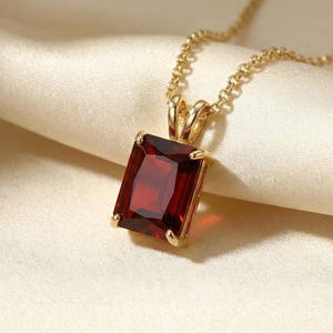 May include: A gold pendant necklace featuring a rectangular, deep red gemstone. The gemstone is set in a gold frame and hangs from a delicate gold chain. The necklace is displayed on a cream-colored, silky fabric.