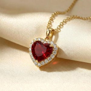 May include: A gold necklace with a heart-shaped pendant. The pendant has a large, deep red gemstone surrounded by a halo of smaller, clear gemstones. The necklace chain is delicate and the pendant is set against a cream-coloured fabric.