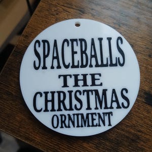 May include: A white, circular Christmas ornament with the text "SPACEBALLS THE CHRISTMAS ORNIMENT" in black, bold letters. The ornament has a small hole at the top for hanging. The design is simple and the text is easy to read.