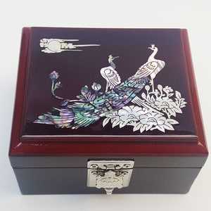 May include: A square jewelry box with a dark red lid and a black base. The lid features an inlaid design of three peacocks and floral elements in iridescent mother-of-pearl. The box has a silver-colored clasp.
