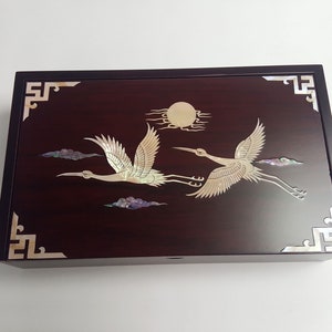 May include: A rectangular, dark wood box with a glossy finish. The lid features an inlaid design of two cranes in flight, a sun, and clouds, all in a pearlescent material. Decorative corner accents complete the design.
