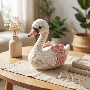 Puede incluir: Un cisne de ganchillo hecho a mano en crema, con detalles rosas y blancos en las alas. El cisne tiene un cuello largo y un pico detallado. Está colocado sobre un camino de mesa de color claro. Un artículo decorativo para una guardería o decoración del hogar.