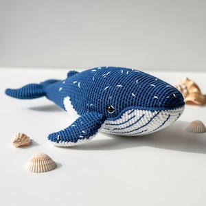 May include: A handmade blue and white crochet whale plush toy with white accents. The whale has a smiling face and is surrounded by seashells. The plush toy is approximately 12 inches long.
