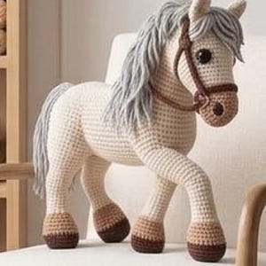 May include: A crocheted horse toy in cream and brown. The horse has a gray mane and tail, brown bridle, and large, black eyes. The toy is standing on a white chair, showcasing its detailed craftsmanship.