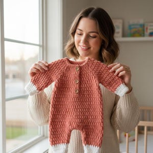 Puede incluir: Un pelele de bebé de ganchillo de color coral con mangas largas y puños y tobillos blancos. El pelele tiene tres botones de madera en la parte delantera. La prenda está hecha a mano y parece suave y cómoda.