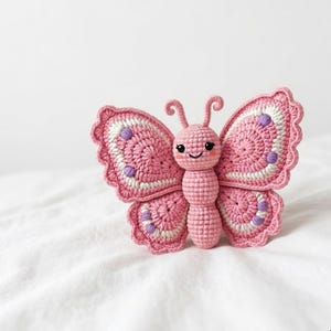 May include: A handcrafted crochet butterfly toy featuring a soft pink body and head with embroidered black eyes and a smiling mouth. The wings are detailed with pink, white, and purple yarn accents, featuring scalloped edges and two curled antennae.