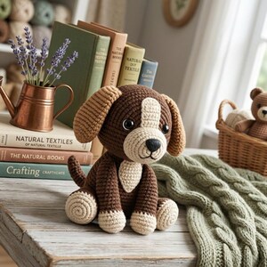 Op de afbeelding: Een bruin en crèmekleurig gehaakt hondenspeeltje met hangende oren, een zwarte neus en donkere ogen. Het speeltje staat voor een stapel boeken met de titels Natural Textiles, The Natural Books en Crafting, naast een koperkleurige kan.