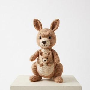 Puede incluir: Un peluche de canguro tejido a ganchillo en tonos marrón y beige. El canguro más grande tiene una bolsa con un canguro más pequeño dentro. El juguete tiene orejas grandes, una cabeza redonda y está sobre una superficie blanca.