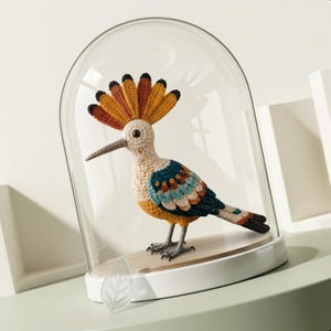 May include: A handcrafted crochet bird with a colorful design, displayed under a glass dome. The bird features a cream-colored body, a long beak, and a vibrant crest of orange, brown, and black feathers. The bird is perched on a wooden base.